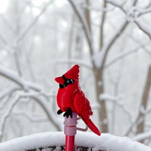 May include: A bright red cardinal bird feeder with a black mask detail, perched on a red pole. The feeder is set against a snowy, wintery backdrop of bare trees and a snow-covered railing.