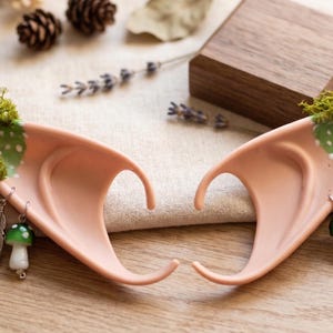 Puede incluir: Un par de orejas de elfo de color melocotón, adornadas con musgo verde, lunares blancos y colgantes. Los colgantes incluyen una flor, una hoja, una pluma y una seta. Perfectas para cosplay o temas de fantasía.