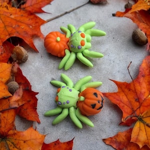 May include: Two green and orange Halloween spider ornaments with pumpkin heads. The spiders have purple dots on their bodies and are sitting on a background of fall leaves.