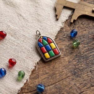May include: A silver-toned pendant shaped like an arched window, featuring inlaid segments of red, blue, green, and yellow enamel. The pendant includes a small metal loop at the top for attachment, displayed alongside loose colorful glass beads.
