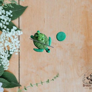 May include: A handmade green turtle-shaped polymer clay charm with a small turtle on its back, a silver pin, and a green circle. The charm is set on a light brown wooden surface, with white flowers and green leaves in the background.