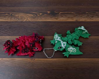 Pode incluir: Duas figuras de dragão feitas à mão. Uma é vermelha com detalhes pretos, a outra verde com bolinhas brancas. Estão conectadas por uma corrente prateada. Os dragões estão sobre uma superfície de madeira castanha escura.