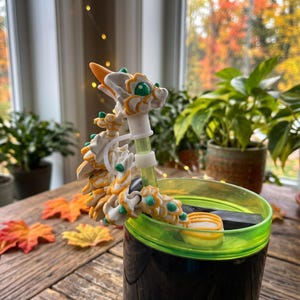 Puede incluir: Un adorno para pajita con forma de dragón articulado en blanco y naranja con detalles en verde, que presenta una cabeza detallada y un cuerpo segmentado. El dragón está sujeto a una pajita verde translúcida en un vaso negro con borde verde.