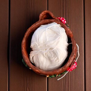 May include: A handmade, brown ceramic bowl filled with a large ball of white yarn. The bowl is decorated with red and white mushroom accents and green leaves. The yarn is a soft, fluffy texture, perfect for knitting or crocheting.