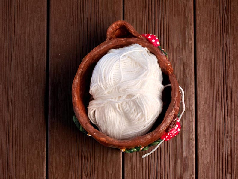 May include: A handmade, brown ceramic bowl filled with a large ball of white yarn. The bowl is decorated with red and white mushroom accents and green leaves. The yarn is a soft, fluffy texture, perfect for knitting or crocheting.
