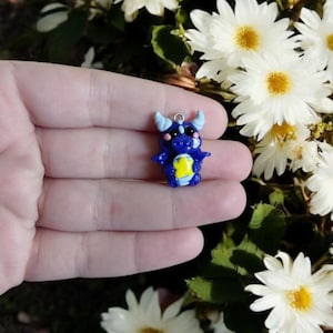 Op de afbeelding: Een kleine blauwe drakenfiguur met lichtblauwe hoorns en vleugels, en een gele ster op de borst. De draak heeft zwarte ogen en een zilverkleurige lus. De achtergrond toont witte bloemen met gele centra.