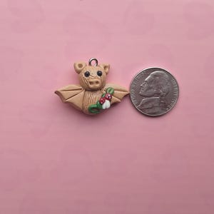 May include: A tan-colored bat-shaped charm with outstretched wings, holding a sprig of red and white berries. The charm has a small metal loop for hanging. A US nickel coin is next to the charm for size comparison. The background is a solid pink color.