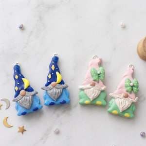 May include: Four gnome charms made of polymer clay. Two gnomes feature dark blue hats with yellow stars and moons, while two gnomes feature light pink hats with green polka-dot bows. All gnomes have gray beards and silver metal jump rings at the top.