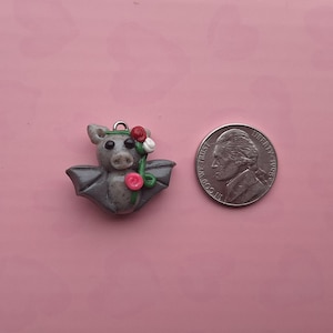 May include: A gray bat charm with a floral design, next to a nickel for size comparison. The bat has gray wings and a small flower crown. A stem with red and white roses is held in its arms. The charm has a silver loop for hanging.