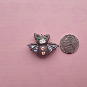 May include: A gray bat-shaped charm with green eyes, red accents, and blue wing details. The charm is next to a US nickel coin on a pink background. The bat has a small metal loop for hanging.