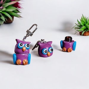 May include: Three purple owl-shaped keychains with blue wings and eyes. One owl has a small glass bottle inside and is not attached to a keychain.