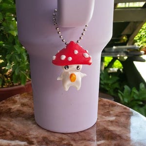 May include: A white and red mushroom charm with a white face and orange eyes. The charm is attached to a silver chain.