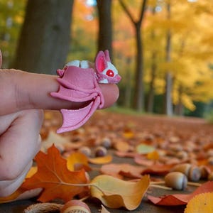Puede incluir: Una pequeña figura de murciélago rosa con detalles blancos y ojos rojos. El murciélago, con las alas extendidas, está posado sobre un dedo. El fondo muestra hojas otoñales en tonos amarillos y naranjas.