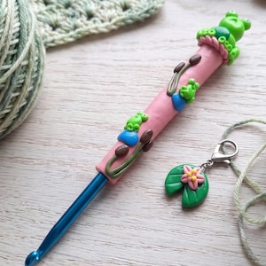 May include: A pink polymer clay crochet hook handle decorated with small green frogs, blue lily pads, and brown cattails, featuring a metallic blue hook tip and a detachable stitch marker charm shaped like a pink flower on a green lily pad.