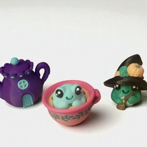 May include: A whimsical collection of miniature polymer clay figurines. Includes a purple teapot, a frog in a pink teacup, and a green frog wearing a witch hat with a pumpkin. The figurines have cute, cartoonish features.