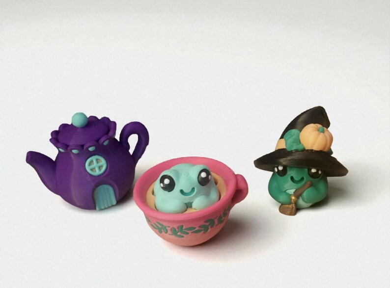 May include: A whimsical collection of miniature polymer clay figurines. Includes a purple teapot, a frog in a pink teacup, and a green frog wearing a witch hat with a pumpkin. The figurines have cute, cartoonish features.