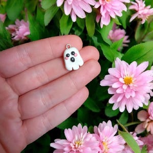 May include: A small white ghost charm with black eyes and pink cheeks. The charm is hanging from a silver loop.