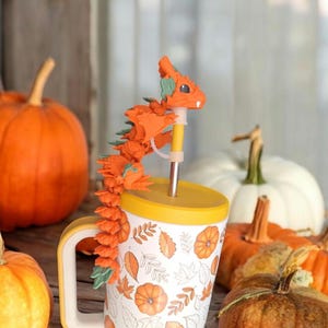 Könnte beinhalten: Ein weißer Becher mit gelbem Rand und einem Herbstlaubmuster, darauf ein orangefarbener und grüner 3D-gedruckter Drachen-Strohhalm-Topper. Der Becher ist von orangefarbenen Kürbissen umgeben.