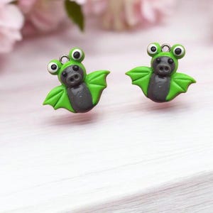 May include: A pair of green and gray bat earrings with googly eyes. The bats are made of polymer clay and have a cute, cartoonish style.