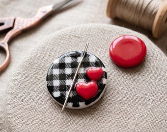 May include: A circular needle minder featuring a black and white gingham pattern with two small red heart-shaped embellishments. A silver metal sewing needle is positioned diagonally across the surface. A separate solid red circular button is nearby.
