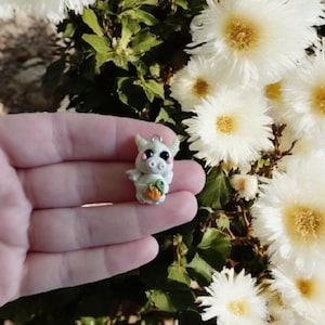 May include: A small, handmade, light gray pig figurine with a pumpkin. The pig has pink cheeks, black eyes, and small wings. It is held in a hand against a backdrop of white flowers and green foliage.