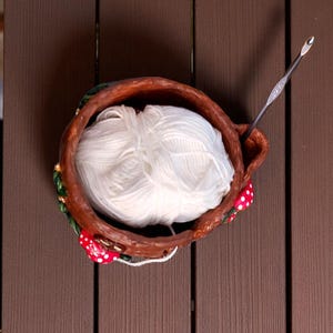 May include: A handmade ceramic yarn bowl with a ball of white yarn and a silver crochet hook. The bowl is brown with red and white mushroom accents and green leaves. The yarn is a soft, fluffy texture.