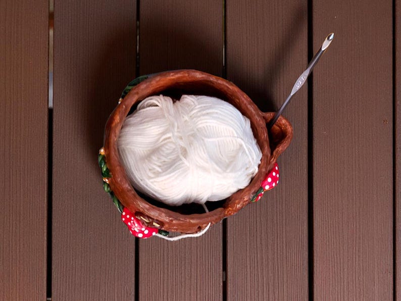 May include: A handmade ceramic yarn bowl with a ball of white yarn and a silver crochet hook. The bowl is brown with red and white mushroom accents and green leaves. The yarn is a soft, fluffy texture.