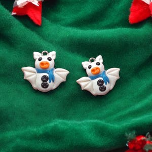 May include: Two white bat ornaments with orange snouts, blue scarves, and black button eyes. The ornaments are on a green fabric background.
