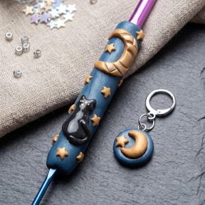 May include: A purple crochet hook with a blue handle decorated with a sculpted black cat, gold crescent moon, and gold stars. A matching circular stitch marker features a gold crescent moon and star on a blue background, completing the set.