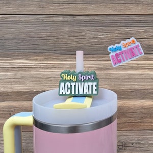 May include: A pink travel mug with a yellow lid and handle. A green and yellow sticker with the text "Holy Spirit Activate" is attached to the lid.