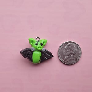May include: A green bat-shaped charm with black glitter wings and a silver loop for hanging. The bat is next to a nickel coin on a pink background. The charm has a stitched design on its belly.