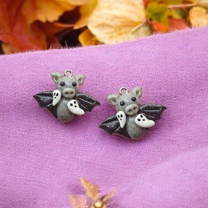 May include: A pair of gray and black bat earrings with white ghost accents. The bats have a cute, cartoon-like appearance.
