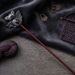 May include: A long, thin, dark red knitting needle featuring a sculpted gray bat topper with glittery black wings, small pink accents, and a central black gemstone. The needle is positioned alongside a dark purple yarn skein and a knitted swatch.