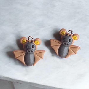 May include: Two brown and gray bat charms with yellow eyes. The bats have a small loop at the top for hanging.