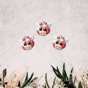 May include: Three white cow charms with red spots and black eyes. The charms have a gold loop for hanging.