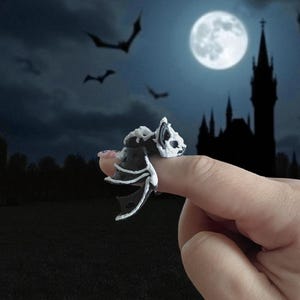 May include: A black and white bat ring with a skeletal design. The ring is shaped like a bat with its wings spread. The background features a full moon, bats, and a silhouette of a castle, creating a Halloween theme.