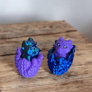 May include: Two small purple and blue dragon figurines, each inside a matching egg-shaped container. The dragon figurines are in a hatching pose with their heads poking out of the eggs.