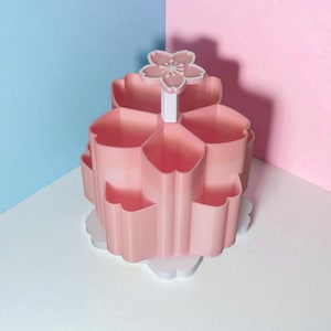 Puede incluir: Un organizador rosa de varios niveles con forma de flor, con compartimentos en forma de pétalo. El organizador tiene una base blanca y un asa en forma de flor. El fondo está dividido entre azul y rosa.