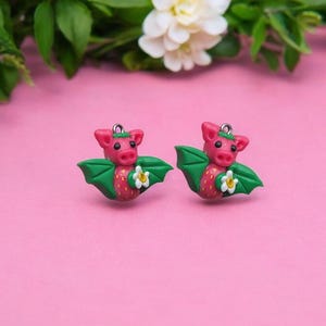 May include: A pair of pink and green bat earrings with a white flower. The bats are made of polymer clay and have a cute, cartoonish style.