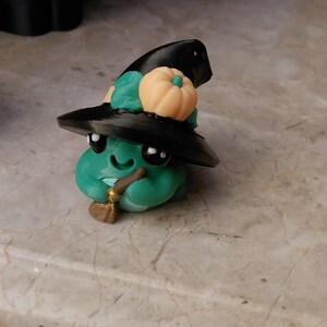 May include: A whimsical green creature figurine wearing a black witch hat adorned with a small orange pumpkin and green leaves. The figurine has large black eyes, a smiling mouth, and holds a small broom and pipe. A Halloween-themed decorative item.