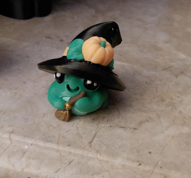 May include: A whimsical green creature figurine wearing a black witch hat adorned with a small orange pumpkin and green leaves. The figurine has large black eyes, a smiling mouth, and holds a small broom and pipe. A Halloween-themed decorative item.