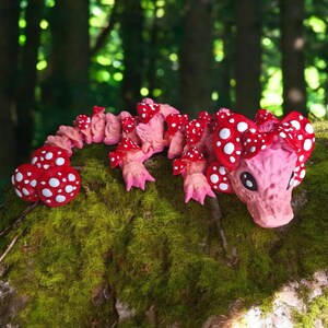 Puede incluir: Una figurita de dragón de color rosa y rojo con forma de hongo y con lunares blancos. El dragón está tumbado sobre un tronco cubierto de musgo.
