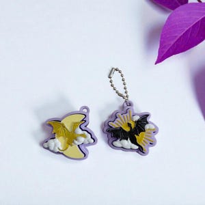 Puede incluir: Dos llaveros morados y amarillos con temática de dragones. Un llavero presenta un dragón amarillo contra una luna amarilla y nubes blancas. El otro llavero tiene un dragón negro con detalles amarillos, rayos de sol y nubes blancas. Una cadena plateada está unida a uno.