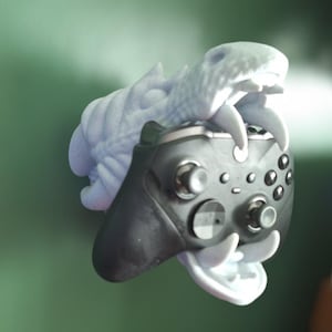 Op de afbeelding: Een 3D-geprinte drakenkop wandbevestiging voor een videogamecontroller. De draak is lichtblauw en heeft een open mond. De controller is zwart en heeft knoppen en een joystick.