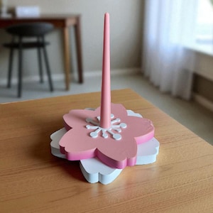 May include: A pink and white wooden floral-shaped paper towel holder. The holder features a tall, slender pink central post and a layered base with a pink flower shape on top of a white flower shape. The holder is on a wooden table.