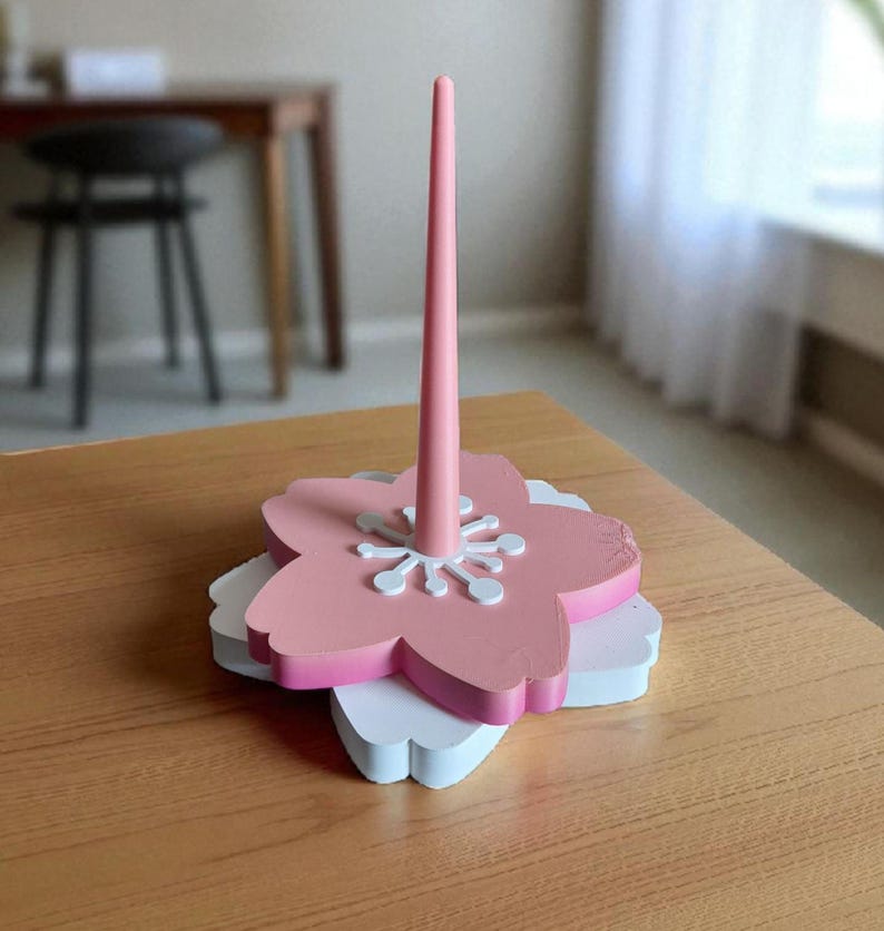 May include: A pink and white wooden floral-shaped paper towel holder. The holder features a tall, slender pink central post and a layered base with a pink flower shape on top of a white flower shape. The holder is on a wooden table.