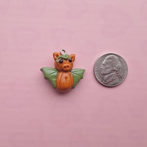 May include: A handmade orange and green bat-shaped charm with a silver loop for hanging. The charm is next to a US nickel coin for size comparison. The background is a solid pink color.