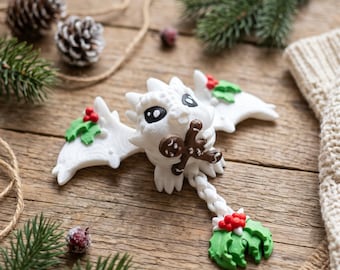May include: A white dragon figurine with large black eyes, holding a small brown gingerbread man. The dragon features white wings decorated with green holly leaves and red berries, and a tail ending in a cluster of green holly and red berries.