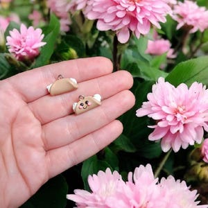 May include: Two small, brown clay taco charms with white details. One taco has a smiling face with pink cheeks. The charms are held in a hand against a background of pink flowers.