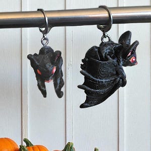 May include: Two black bat-shaped Halloween ornaments with red accents hang from a metal rod. The bats are detailed with textured wings and faces. Several orange pumpkins are in the foreground, suggesting a fall or Halloween theme.
